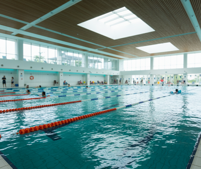 Modern indoor pool in Singapore with turquoise water, glass panels, families swimming, bright orange markers.